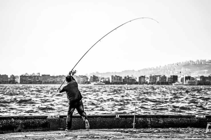 grayscale photography of man holding a fishing rod near body of water